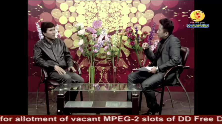 Two men sit in chairs, facing each other on a DD Arun Prabha set, with a table between them. The man on the right gestures as he speaks, while the other listens.
