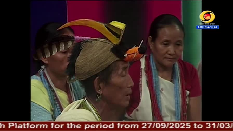 A man wearing a traditional hat with a large yellow beak speaks, his face lit by the studio lights. Two women flank him, one with a scarf and the other with feathers in her hair, as they appear on DD Arun Prabha.
