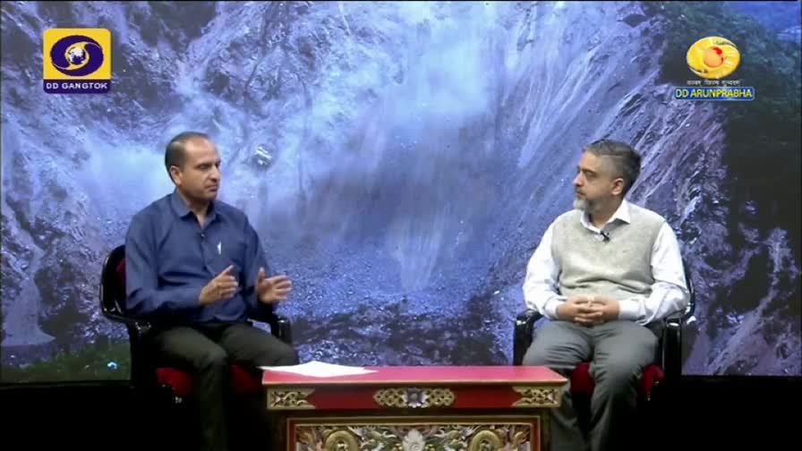 Two men sit in chairs, facing each other on a set for DD Arun Prabha, the background a painted landscape. The man on the left gestures with his hands while speaking, while the other listens intently.
