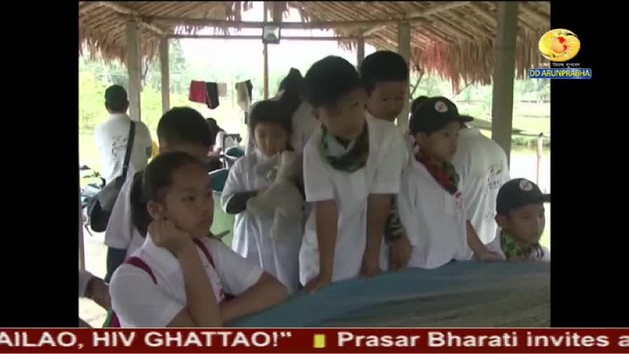 Children in white shirts and caps lean forward, gazing at something just out of view. A DD Arun Prabha logo sits in the corner, as they appear to be watching something on the water.
