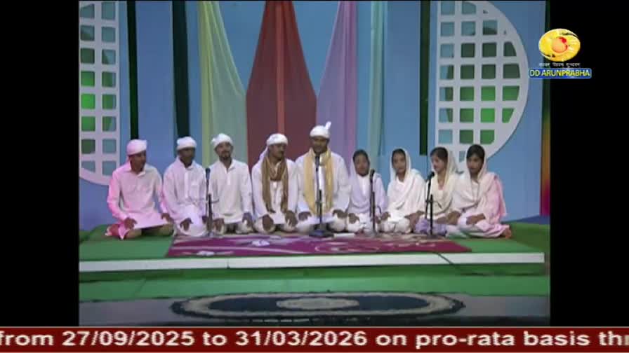 A group of people in white clothing are seated on a stage, facing forward. Microphones stand before them, and the DD Arun Prabha logo is visible in the corner.
