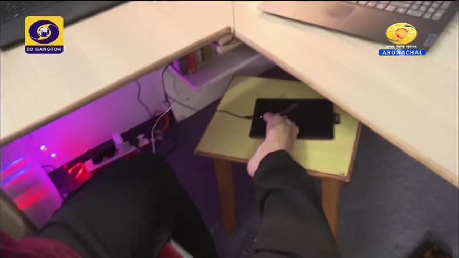 Beneath the desk, a person's foot rests on a small table, a stylus in hand, drawing on a tablet. The logos for DD Gangtok and DD Arun Prabha are visible near the desk's edges.
