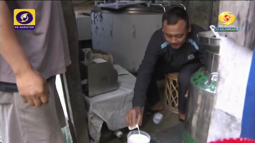 A man, seated on a stool, carefully pours a stream of white liquid from a small container into a clear cup. The scene is likely being filmed for DD Arun Prabha, as the logo is visible in the corner.

