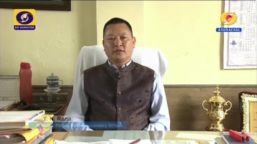 A man in a patterned vest sits behind a desk, speaking directly to the camera, likely for DD Arun Prabha. The logos for DD Gangtok and Arunachal are visible, indicating the broadcast's origin.
