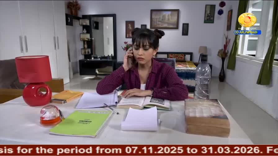 A young woman in a maroon shirt is talking on the phone while studying at a table, books and papers scattered around. The DD Arun Prabha logo is visible in the corner, suggesting this is a scene from an Indian television show.
