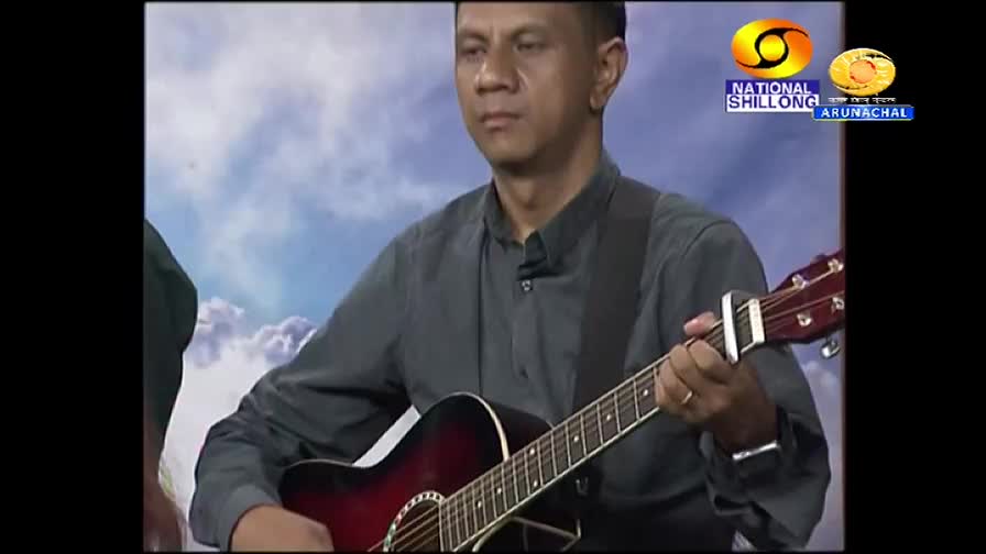 A man in a gray shirt strums a maroon acoustic guitar, fingers moving over the frets.  Behind him, a cloudy sky backdrop and logos for "National Shillong" and DD Arun Prabha, India, are visible.
