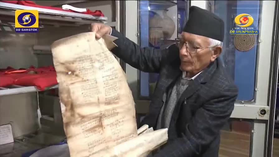 An older man, wearing a black cap, carefully holds up a large, aged document, its surface covered in faded script. The DD Arun Prabha logo is visible as he displays the fragile paper.
