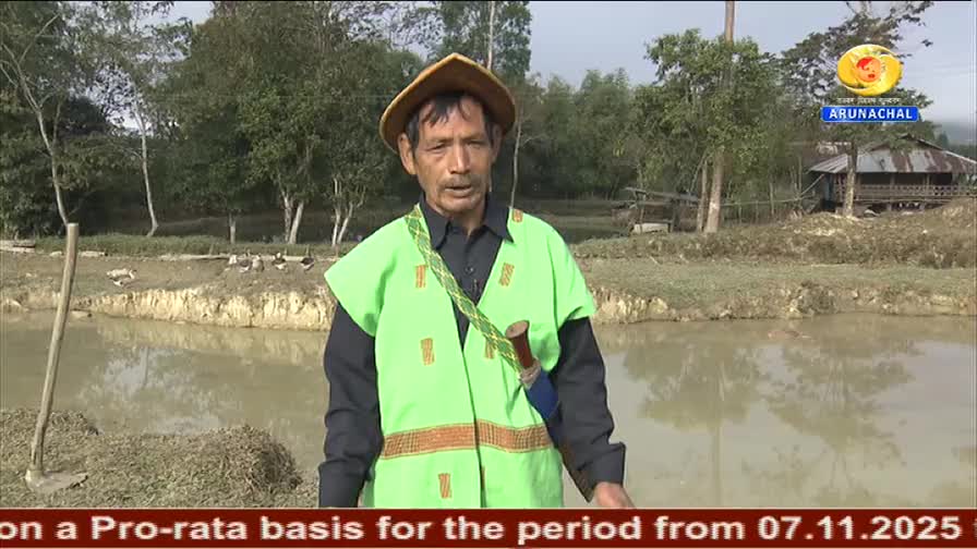 The man, wearing a green vest and hat, is speaking near a body of water. Behind him, a DD Arun Prabha logo is visible, and a caption scrolls across the bottom of the screen.
