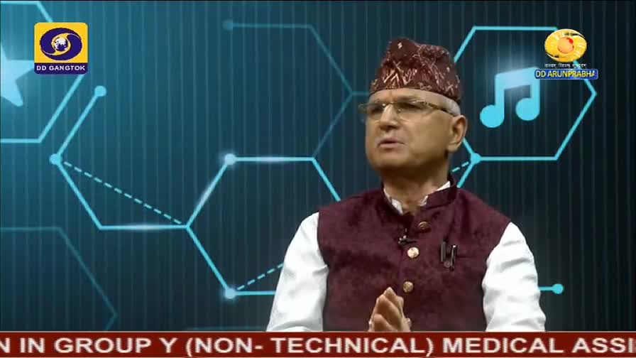 A man in a patterned cap and glasses gestures with his hands, speaking on a set for DD Arun Prabha. The background features a blue graphic with the logos for DD Gangtok and a musical note.
