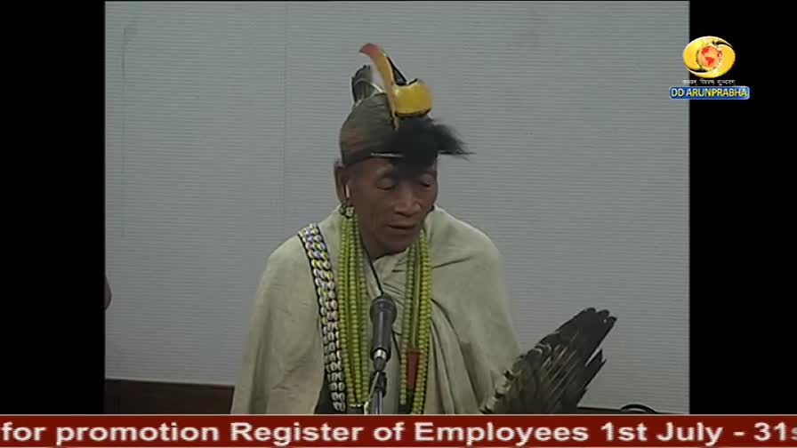 A man in a feathered headdress and beaded necklace speaks into a microphone, likely on DD Arun Prabha. He holds a large fan in his right hand.
