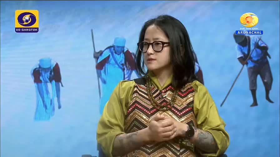A woman in glasses and a patterned top is speaking, her hands clasped in front of her. Behind her, stylized figures in blue and white are depicted against a light blue backdrop, likely on a DD Arun Prabha broadcast from India.
