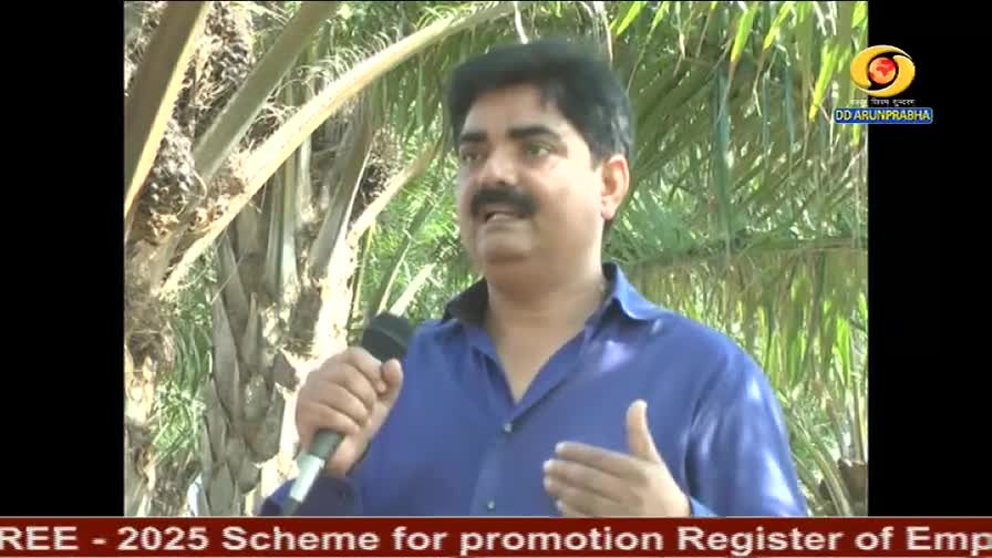 A man in a blue shirt holds a microphone, gesturing as he speaks. The DD Arun Prabha logo is visible, and palm trees are behind him.
A man in a blue shirt holds a microphone, gesturing as he speaks. The DD Arun Prabha logo is visible, and palm trees are behind him.