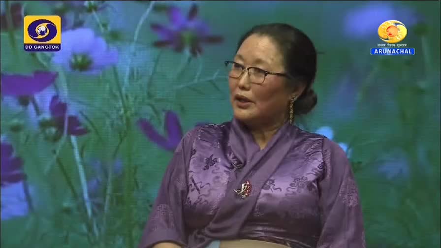 A woman in a purple robe speaks, framed by a floral backdrop, with the DD Gangtok and Arunachal Pradesh logos visible. She gestures with her hands as she speaks, likely on the DD Arun Prabha channel.
A woman in a purple robe speaks, framed by a floral backdrop, with the DD Gangtok and Arunachal Pradesh logos visible. She gestures with her hands as she speaks, likely on the DD Arun Prabha channel.