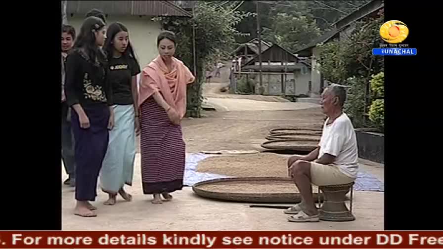 A group of women in traditional dress stand facing an older man seated on a stool, with drying grains spread out on large, round trays nearby. The scene appears to be part of a DD Arun Prabha broadcast, with a logo in the corner.
A group of women in traditional dress stand facing an older man seated on a stool, with drying grains spread out on large, round trays nearby. The scene appears to be part of a DD Arun Prabha broadcast, with a logo in the corner.
