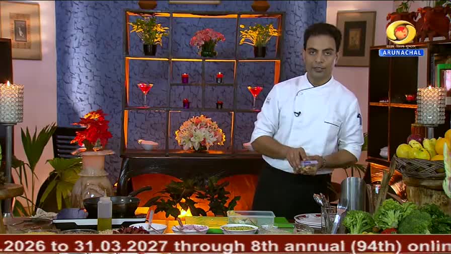 A chef in a white uniform stands before a backdrop of shelves adorned with flowers and candles, holding a small container of nuts. On the counter in front of him, ingredients like broccoli and lemons are arranged, hinting at a cooking demonstration for DD Arun Prabha. A chef in a white uniform stands before a backdrop of shelves adorned with flowers and candles, holding a small container of nuts. On the counter in front of him, ingredients like broccoli and lemons are arranged, hinting at a cooking demonstration for DD Arun Prabha.