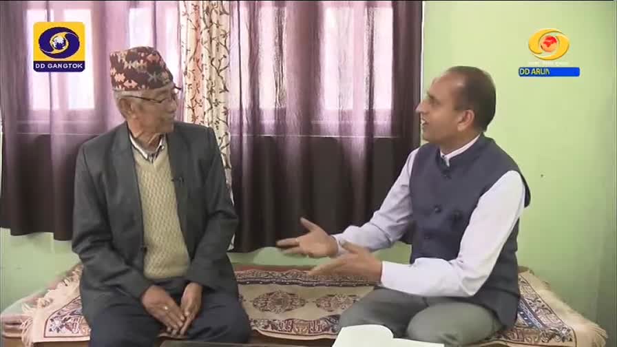 Two men sit on a sofa, engaged in conversation. The man on the left wears a traditional cap and a suit jacket over a patterned sweater. The man on the right, in a dark vest over a light shirt, gestures animatedly. A DD Gangtok logo is visible in the upper left corner. Two men sit on a sofa, engaged in conversation. The man on the left wears a traditional cap and a suit jacket over a patterned sweater. The man on the right, in a dark vest over a light shirt, gestures animatedly. A DD Gangtok logo is visible in the upper left corner.