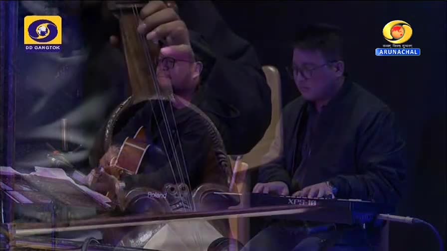 A musician plays a keyboard, his fingers moving across the keys. Beside him, another man draws a bow across the strings of a large stringed instrument. The DD Arun Prabha logo for Arunachal Pradesh is visible. A musician plays a keyboard, his fingers moving across the keys. Beside him, another man draws a bow across the strings of a large stringed instrument. The DD Arun Prabha logo for Arunachal Pradesh is visible.