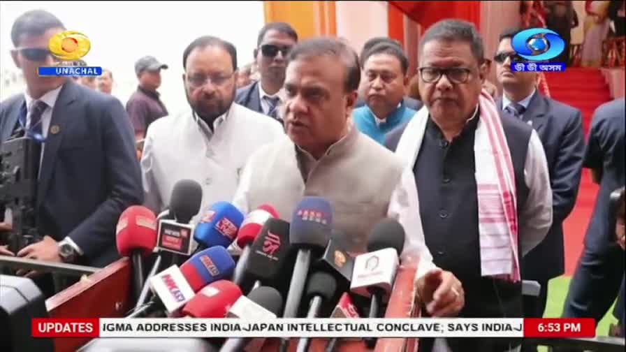 A man in a light-colored vest speaks into a cluster of microphones, his expression earnest. Behind him, a man in a dark suit and glasses stands with a white and red striped scarf draped over his shoulders.
