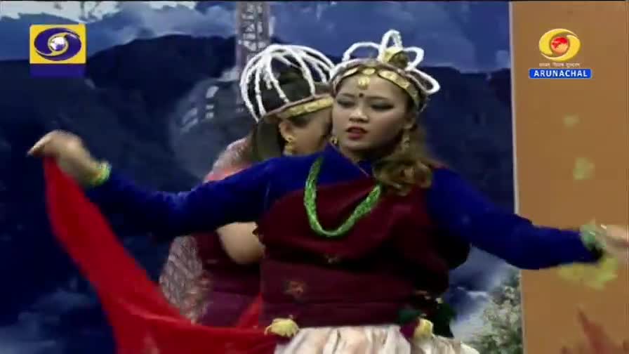 A dancer in a blue and maroon outfit gracefully extends a flowing red fabric. Another performer, adorned with a white headdress, stands behind her, also in traditional attire.