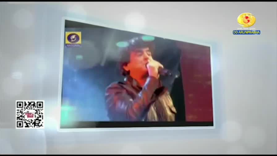 A man in a dark jacket sings into a microphone on a stage, bathed in blue light. The DD Arun Prabha logo appears in the corner.