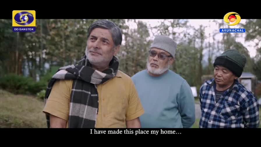 Three men stand together outdoors, their faces turned slightly away from the camera. The man in front wears a yellow shirt and a checkered scarf, while the other two are in muted sweaters and caps. A DD Arun Prabha logo appears in the upper right corner.