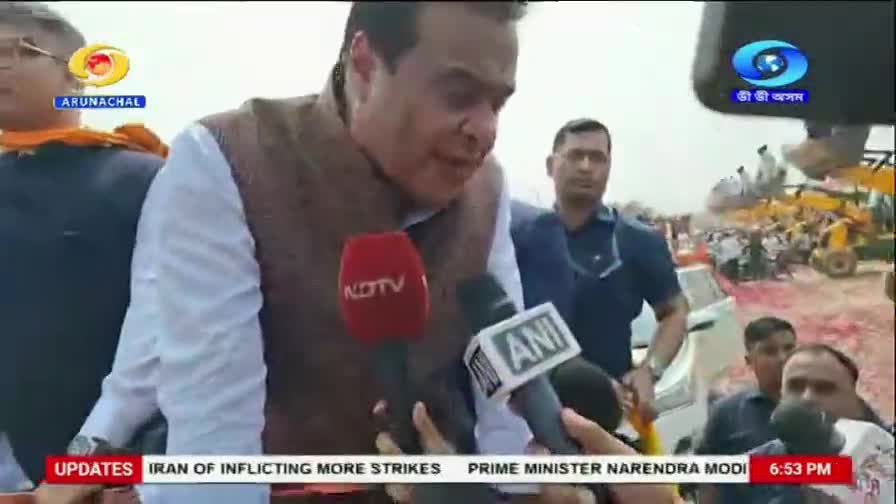 A man in a brown vest speaks into a red NDTV microphone, while an ANI microphone is held close by. Behind him, a crowd gathers near yellow construction equipment.