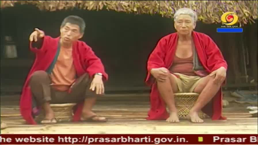 Two men in bright red robes sit on woven stools. The man on the left gestures with his right hand, his mouth open as if speaking. The DD Arun Prabha logo is visible in the upper right corner.