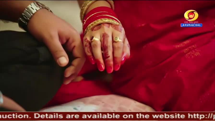 A man's hand rests on a woman's lap, her fingers adorned with red alta and gold bangles. The DD Arun Prabha logo is visible on her bright red sari.