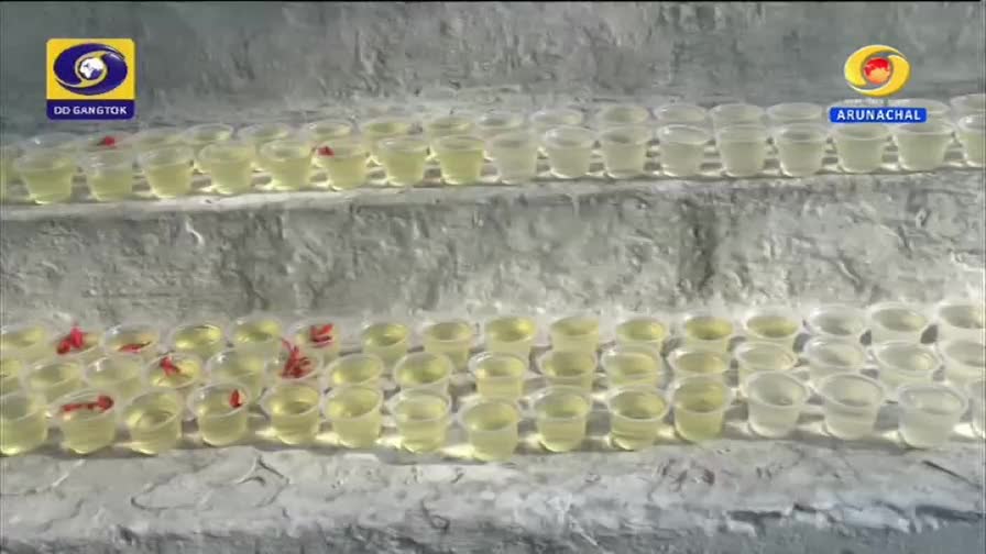 Rows of small plastic cups, each holding a bit of yellow liquid and some with bright red fish, are arranged on tiered steps. The DD Arun Prabha logo is visible in the corner, indicating this scene is from India.