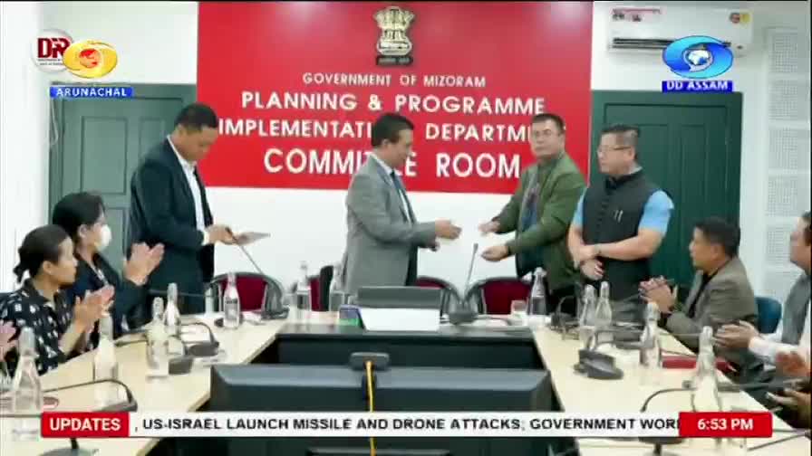 A man in a suit hands a document to another man across a table. People seated around them clap, their hands meeting in mid-air.