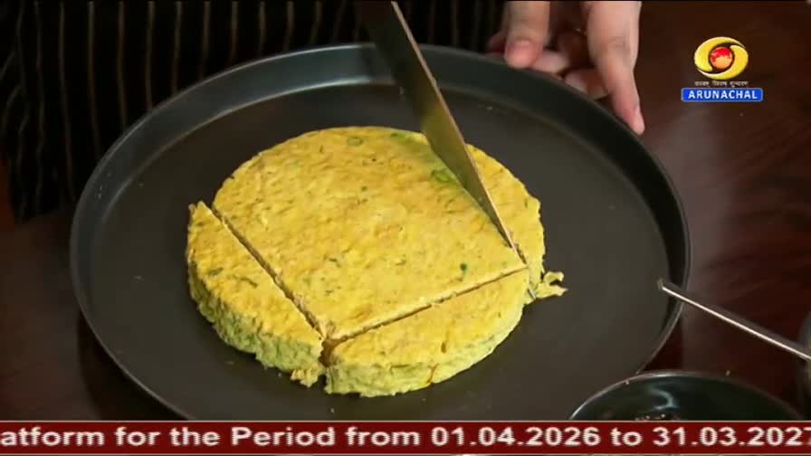 A knife slices through a round, yellow dish on a black pan, dividing it into wedges. This cooking demonstration is part of a DD Arun Prabha broadcast from India.