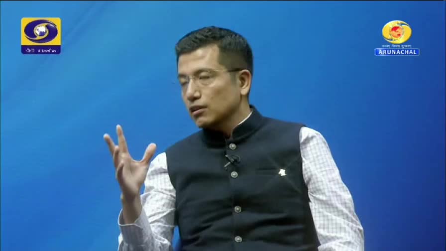 A man in a dark vest over a patterned shirt speaks with his hand raised, gesturing. The DD Arun Prabha logo is visible on the left, and the Arunachal logo on the right. A man in a dark vest over a patterned shirt speaks with his hand raised, gesturing. The DD Arun Prabha logo is visible on the left, and the Arunachal logo on the right.