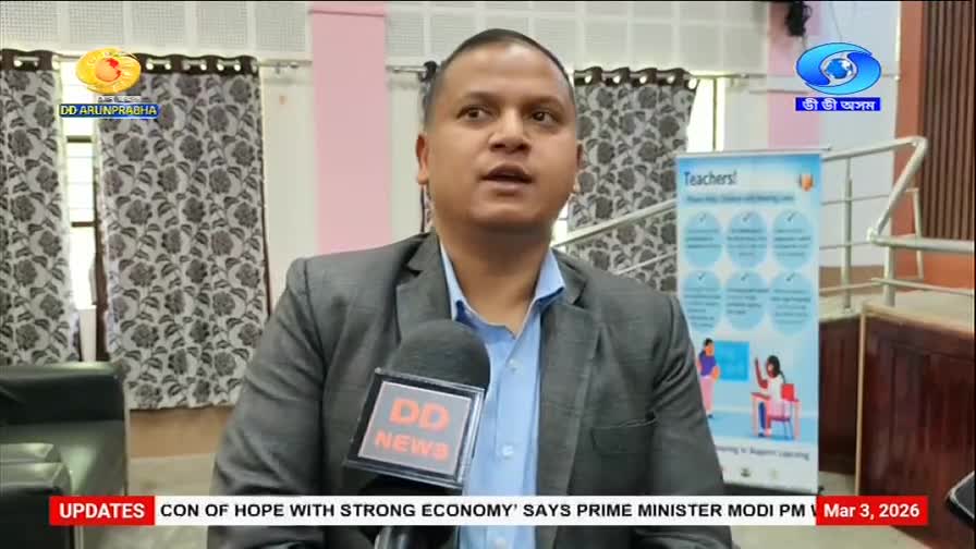 A man in a suit speaks into a DD News microphone, with the DD Arun Prabha logo visible. Behind him, a banner for "Teachers!" and a staircase are present. A man in a suit speaks into a DD News microphone, with the DD Arun Prabha logo visible. Behind him, a banner for "Teachers!" and a staircase are present.