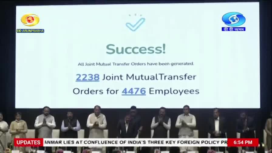 A large screen displays "Success!" with the DD Arun Prabha logo. Below, a group of people in formal attire stand on a stage, some with hands clasped in greeting.
