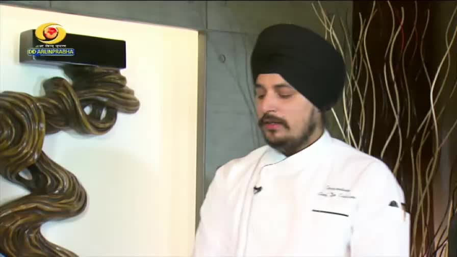 A chef in a white coat and black turban stands ready, a DD Arun Prabha logo visible on the wall behind him. He is positioned next to a decorative, dark brown, twisted sculpture.