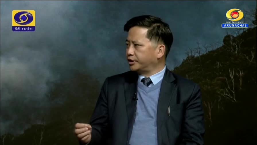 A man in a suit and tie speaks animatedly, his hand gesturing. The DD Arun Prabha logo is visible in the corner, alongside a backdrop of misty, mountainous terrain.