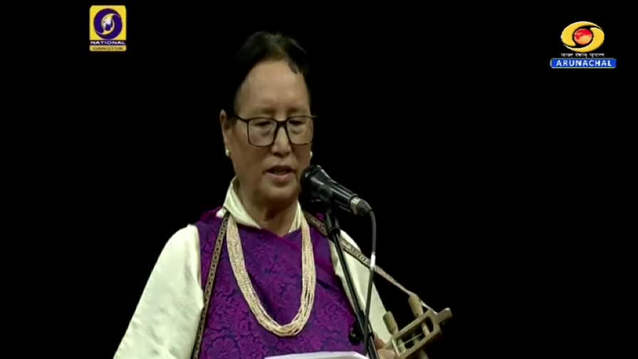 A woman in traditional attire sings into a microphone, holding a small stringed instrument. The DD Arun Prabha logo is visible in the corner.