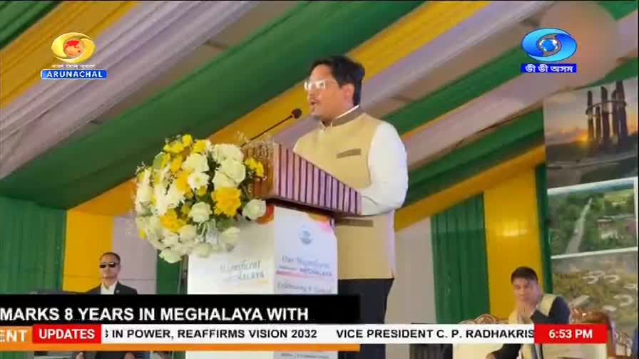 A man in a tan vest speaks at a podium adorned with flowers. The backdrop is a colorful tent, and the DD Arun Prabha logo is visible.