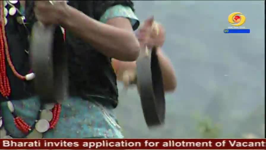 A person in traditional attire, adorned with red beads and silver discs, strikes two metal cymbals together. The DD Arun Prabha logo is visible in the upper right corner.