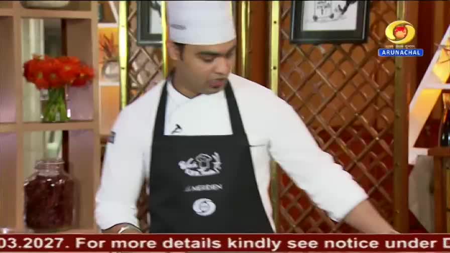 A chef in a white uniform and black apron is demonstrating something in a studio setting, likely for DD Arun Prabha. A jar of red preserves and a bouquet of orange flowers sit on a shelf nearby.