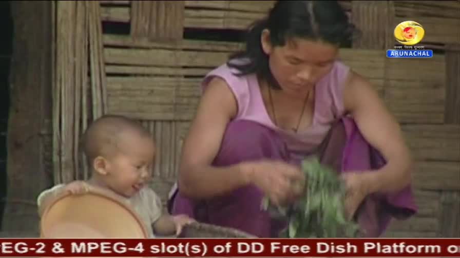A woman in a pink top and purple skirt sorts through a bunch of green leaves, her hands busy with the task. Beside her, a young child with a shaved head leans forward, gripping a light brown bowl. The scene unfolds against a backdrop of woven bamboo walls, a common sight in rural India, as a DD Arun Prabha broadcast plays.
