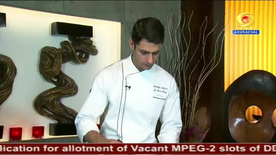 A chef in a crisp white uniform stands before a backdrop of artistic sculptures and dried branches. The DD Arunachal logo is visible in the upper right corner, indicating a broadcast from India.