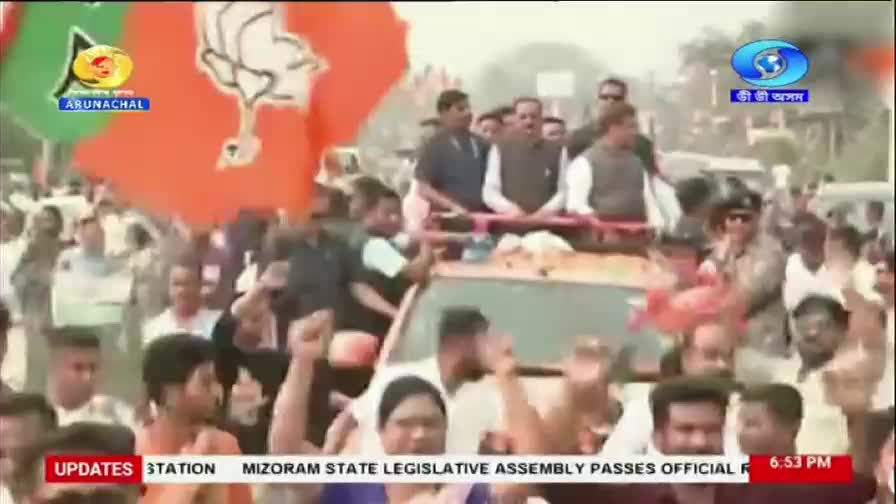 A crowd surges forward, waving large orange and green flags. Men in the back of a vehicle, likely part of a political procession, acknowledge the cheers.