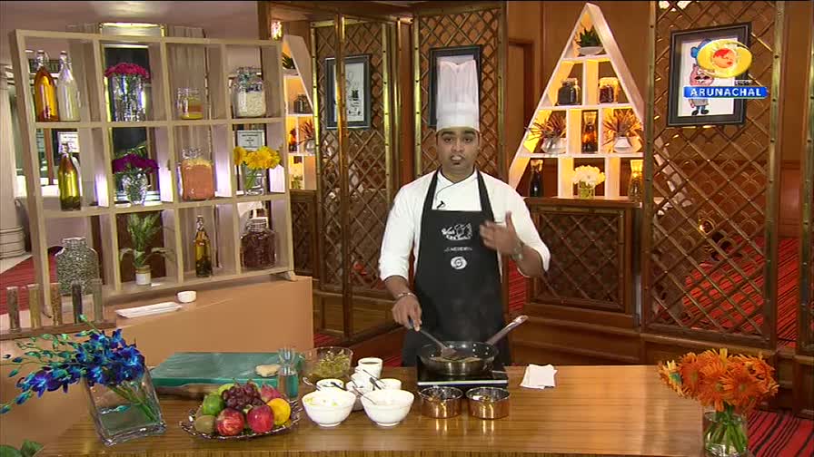 A chef, wearing a tall white hat and a black apron, gestures with his right hand while stirring something in a pan on a small electric stove. Behind him, a shelf unit displays various bottles and jars, and a triangular structure holds more decorative items. The DD Arun Prabha logo is visible in the upper right corner.