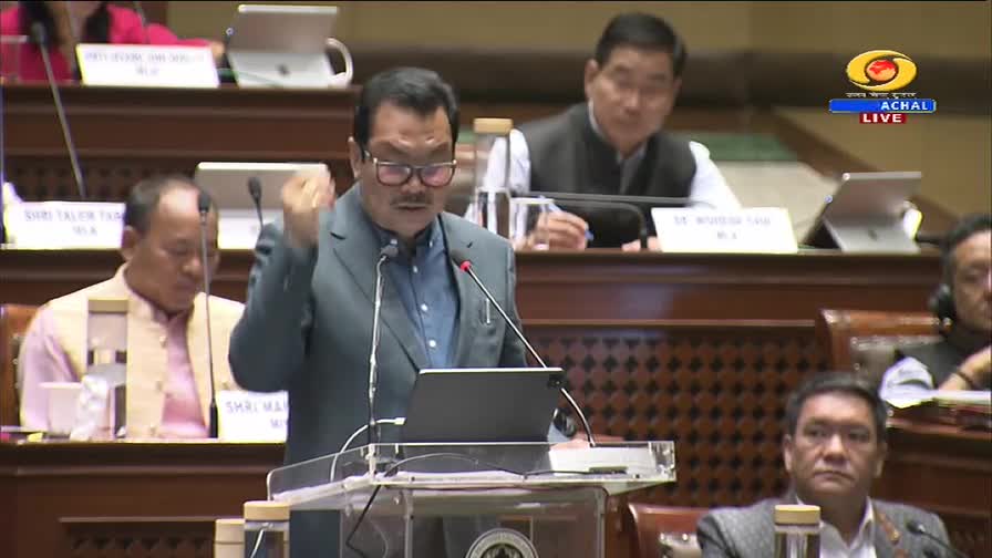 A man in a grey suit speaks animatedly at a podium, his right hand raised. This live broadcast from DD Arun Prabha captures a moment in an Indian legislative session.