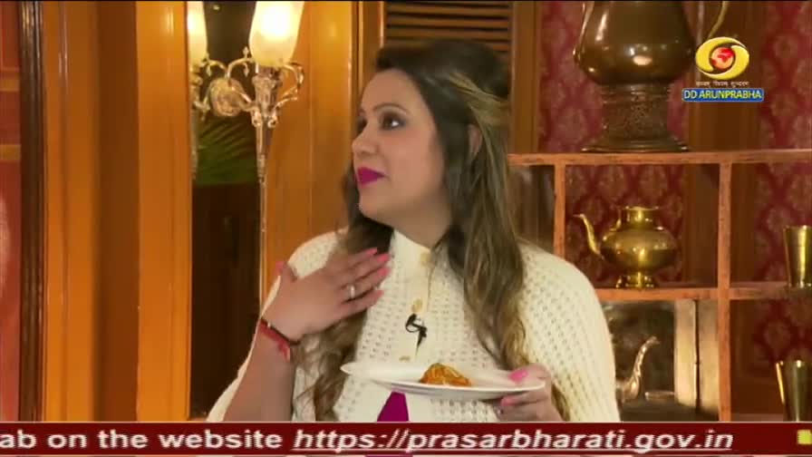 A woman on a DD Arun Prabha broadcast holds a small plate with food. She gestures with her left hand while looking off-camera.