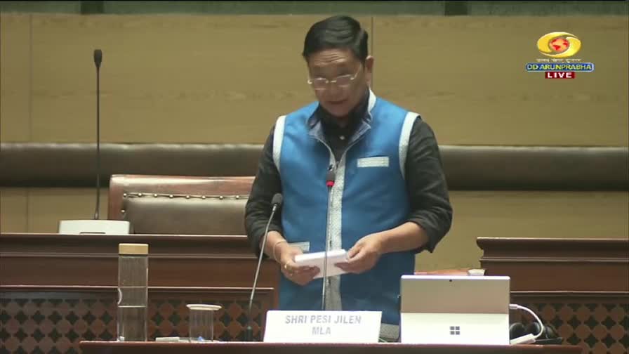 Shri Pesi Jilen, MLA, stands at a podium, holding papers. The DD Arun Prabha live broadcast captures this moment from within the assembly.