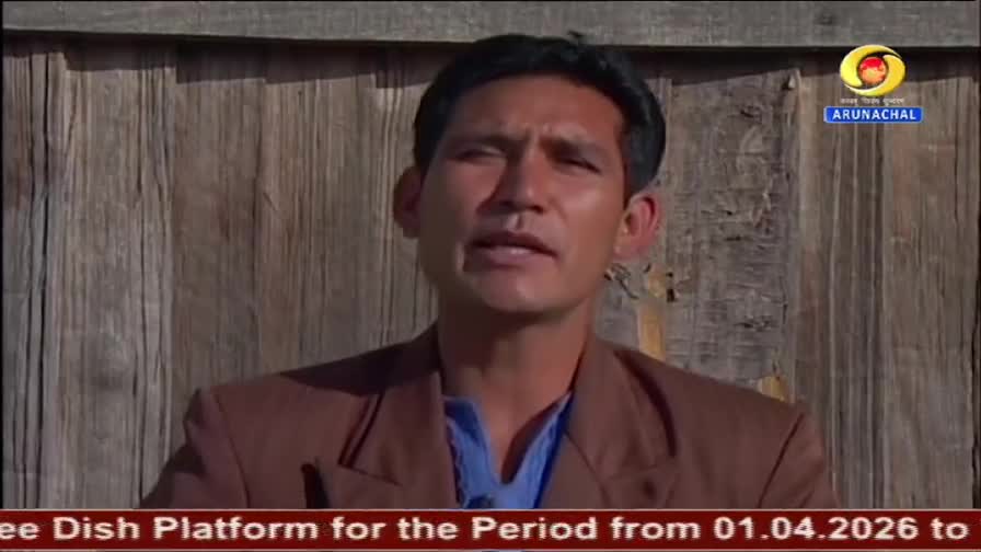 A man in a brown jacket speaks in front of a wooden wall, with the DD Arun Prabha logo visible. A red banner at the bottom displays text about a free dish platform.