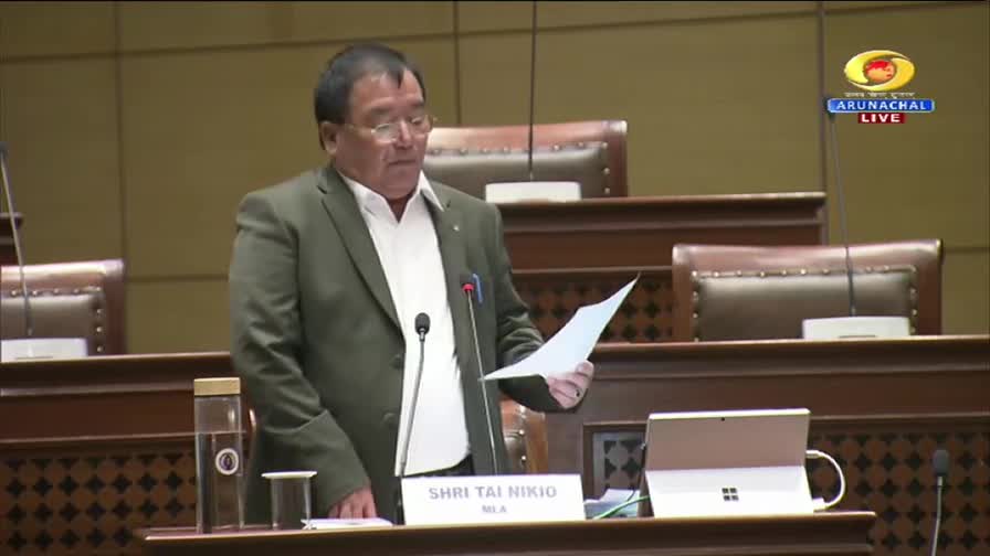 Shri Tai Nikio, MLA, stands at the podium, holding a document. The DD Arun Prabha logo is visible in the corner, indicating this is a live broadcast from Arunachal Pradesh.