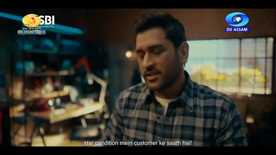 A man in a plaid shirt speaks directly to the camera, his expression earnest. Behind him, a workbench is cluttered with tools and equipment, hinting at a workshop setting.